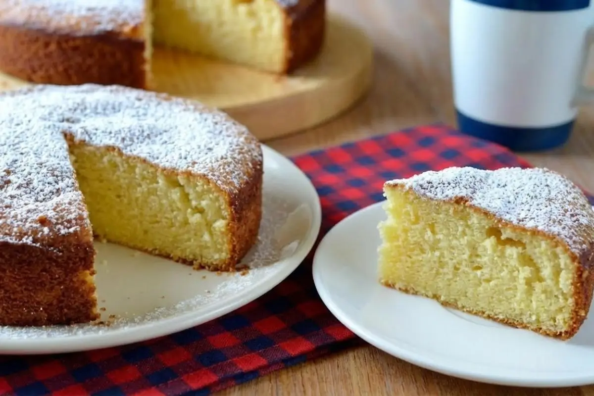 Rena's No-Fail Tips For Your Italian Lemon Ricotta Cake A beautifully sliced Italian lemon ricotta cake dusted with powdered sugar sits on a white plate over a rustic plaid napkin, next to a perfectly plated, moist slice ready to be enjoyed.