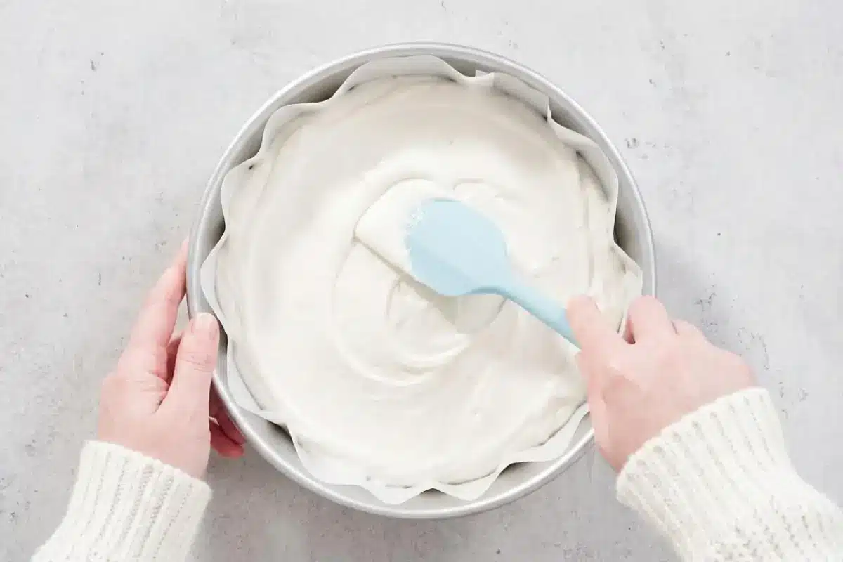 Rena's No-Fail Tips For Your White Almond Cake An overhead view of a baker wearing a cozy sweater using a light blue silicone spatula to gently smooth thick white almond cake batter into a round metal baking pan lined with parchment paper.