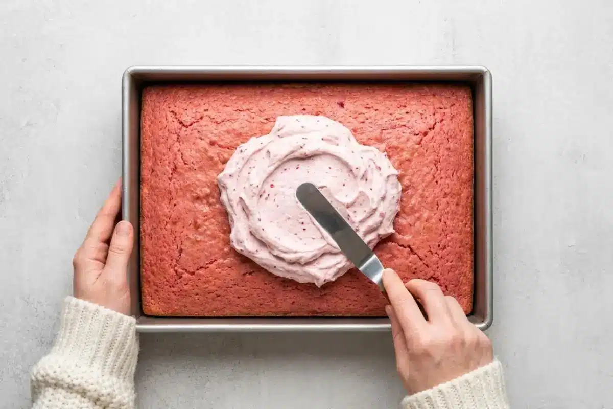 Rena's Secrets for the Perfect Bake For Raspberry Cake A top-down view of a baker wearing a cozy cream sweater, using an offset spatula to spread vibrant pink raspberry frosting across the top of a cooled, rectangular baked raspberry sheet cake inside a metal baking pan.