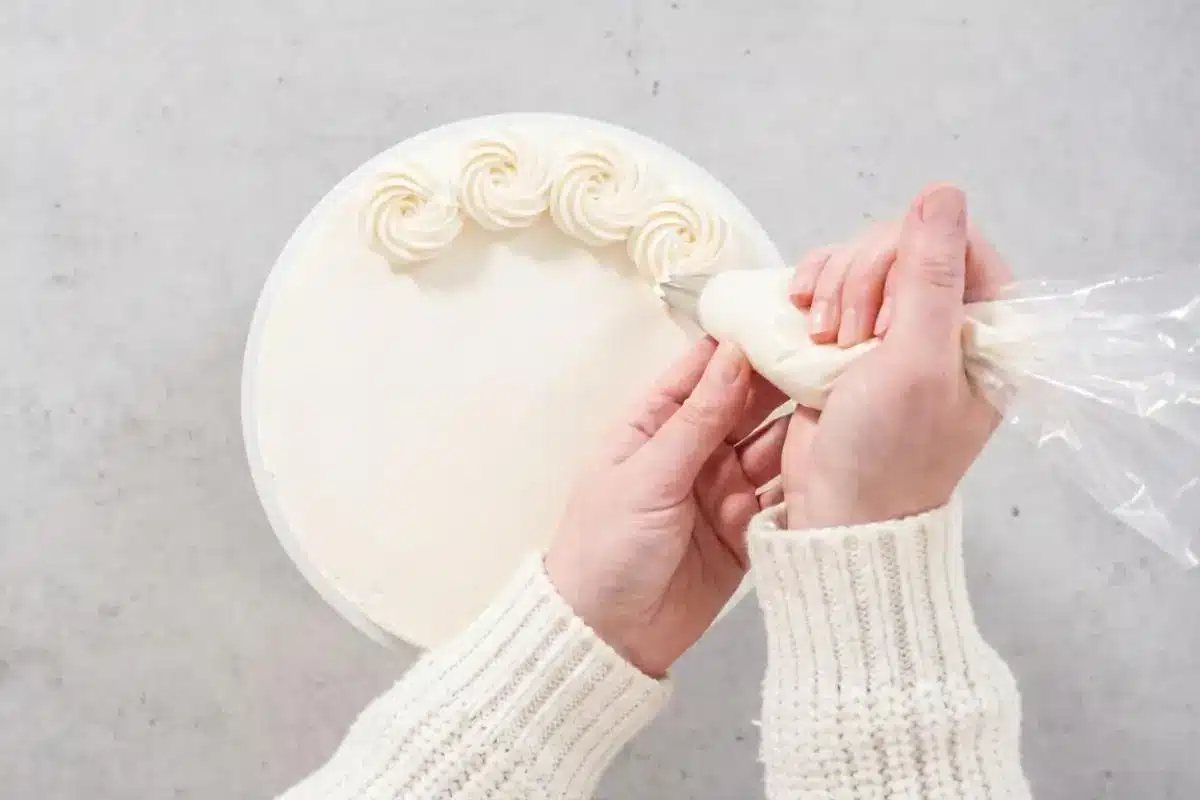 Rena's for Your White Almond Cake An overhead view of a baker's hands in a cozy ribbed sweater using a clear piping bag to gently pipe a border of white buttercream rosettes onto a smooth cake, showcasing no-fail tips for your white almond cake.