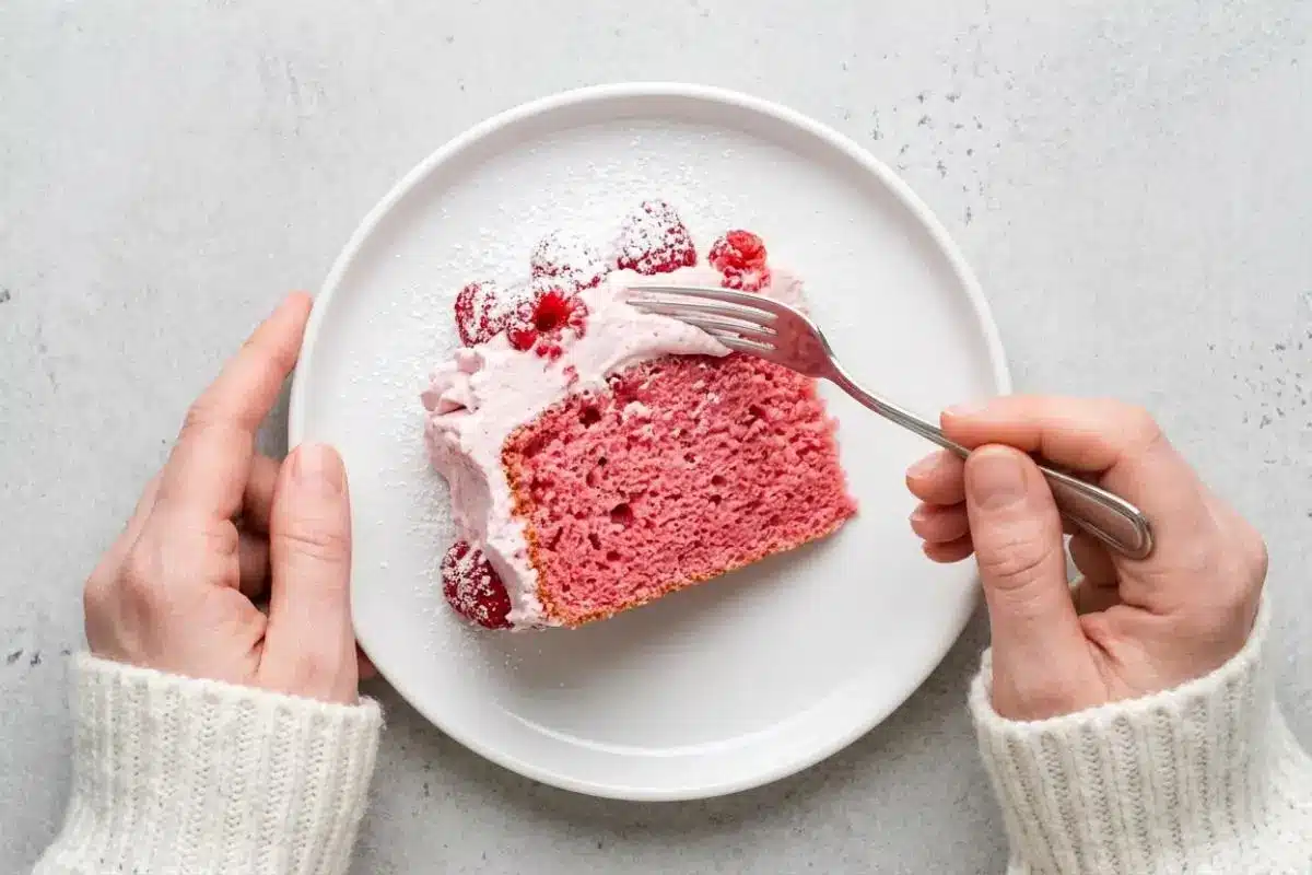 Serving and Storing For Raspberry Cake An overhead view of a person wearing a cozy cream sweater using a silver fork to cut into a freshly served slice of tender pink raspberry cake, topped with creamy frosting and fresh berries on a white plate.