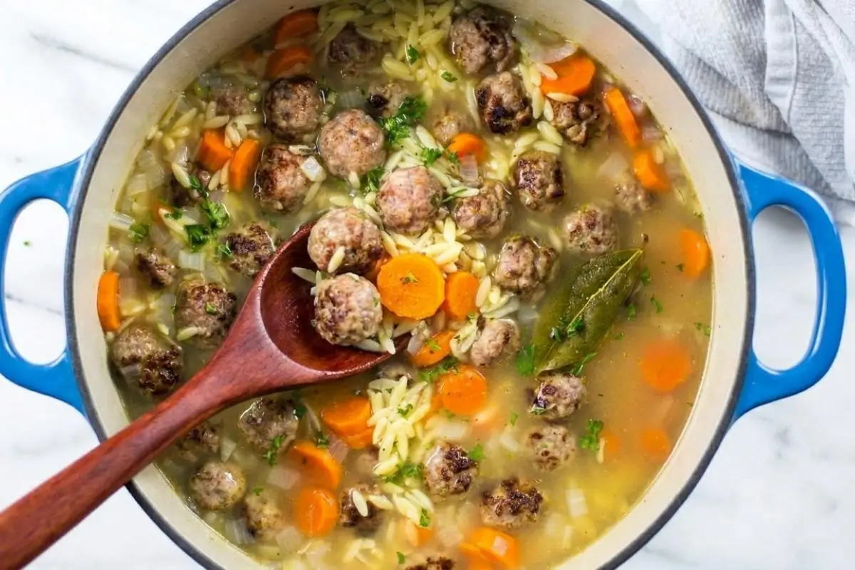 Storage & Freezing Pro-Tip For Your Italian Meatball and Orzo Soup An overhead view of a large blue Dutch oven filled with homemade Italian meatball and orzo soup, featuring tender mini meatballs, sliced carrots, and a bay leaf, with a wooden spoon lifting a hearty scoop from the warm broth.