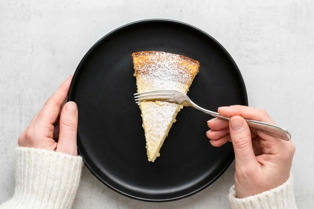 Storage & Freezing Pro-Tip for Limoncello Ricotta Cake A top-down view of a person wearing a cozy cream sweater using a silver fork to cut into a slice of powdered sugar-dusted dessert on a black plate, showcasing the perfectly moist crumb for tips on storing and freezing limoncello ricotta cake.