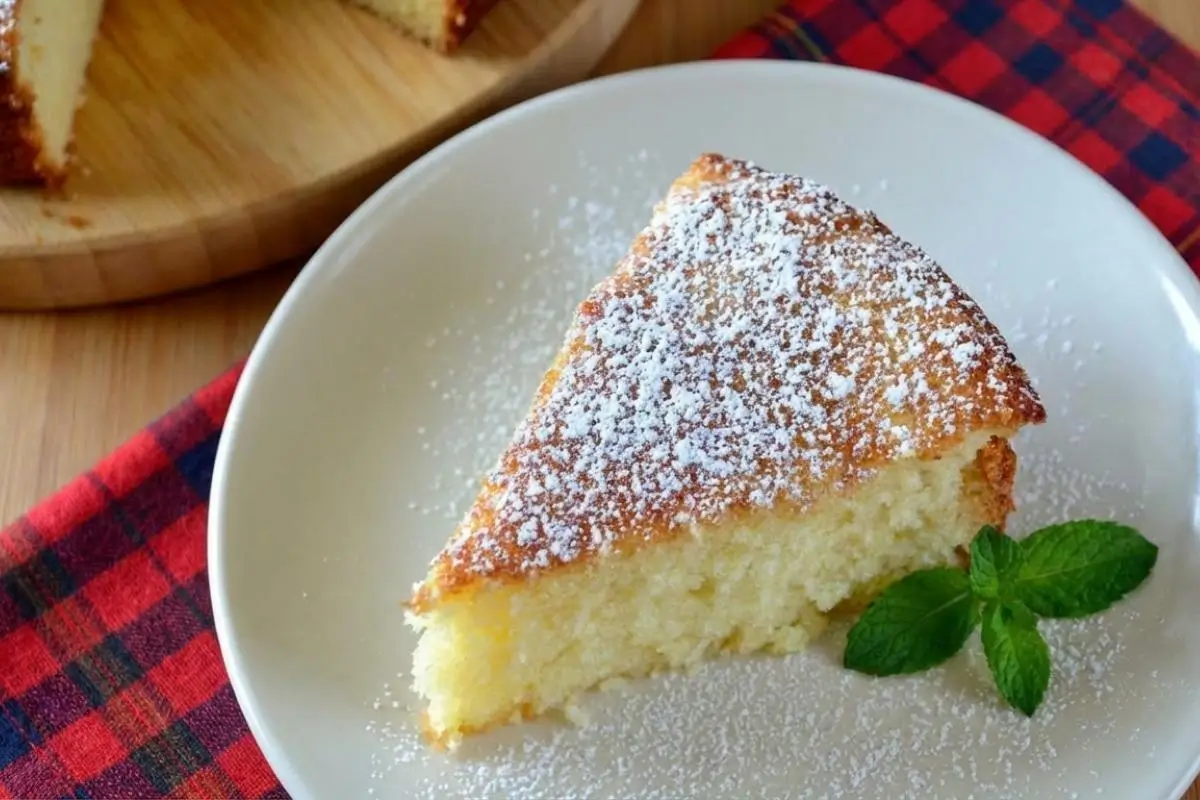 Storage & Freezing Pro-Tip for Your Italian Lemon Ricotta Cake A single slice of moist Italian lemon ricotta cake sprinkled with powdered sugar and garnished with a fresh mint leaf on a white plate, with the remaining cake on a wooden board in the background.