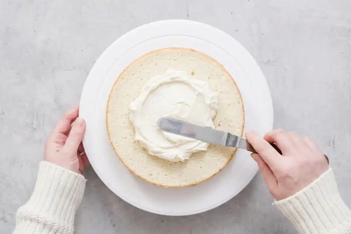 Storage & Freezing Pro-Tip for Your White Almond Cake An overhead view of a baker's hands in a cozy ribbed sweater using a metal offset spatula to gently spread fluffy white buttercream frosting onto a baked layer of white almond cake resting on a white turntable.