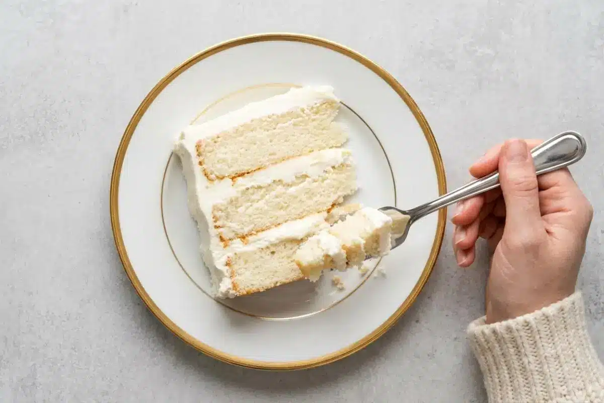 Storage for Your White Almond Cake An overhead view of a hand using a silver fork to take a bite from a moist three-layer slice of cake on a gold-rimmed plate, demonstrating the soft texture preserved by proper storage for your white almond cake.