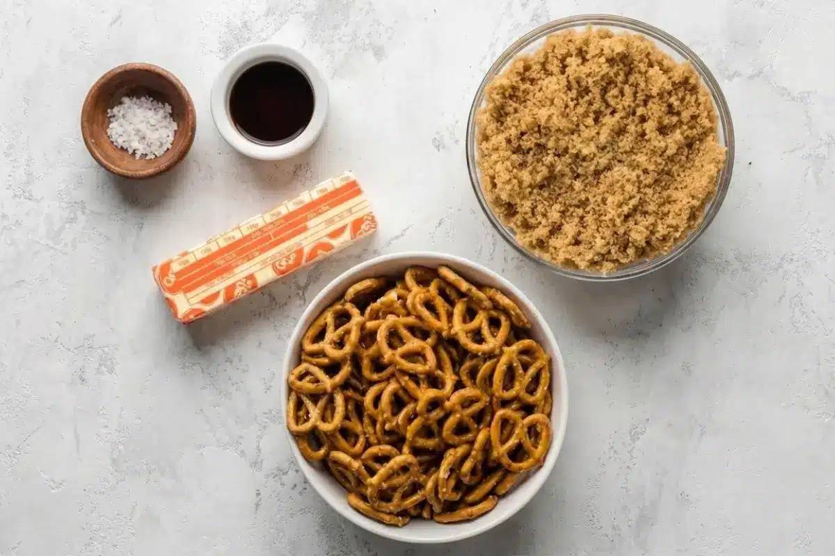 The Foolproof Ingredients List For Your Butter Toffee Pretzel An overhead view of the simple butter toffee pretzel ingredients, featuring a white bowl filled with mini pretzels, a glass bowl of brown sugar, a stick of butter, vanilla extract, and a pinch of flaky sea salt resting on a light, textured surface.