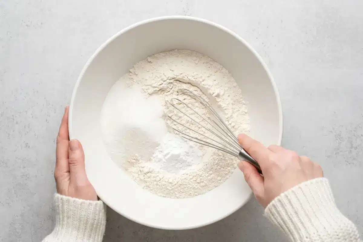 Whisk the Dry Ingredients For Raspberry Cake An overhead view of a baker wearing a cozy cream sweater, using a wire whisk to blend the flour, sugar, and other dry ingredients for raspberry cake in a large white ceramic bowl.