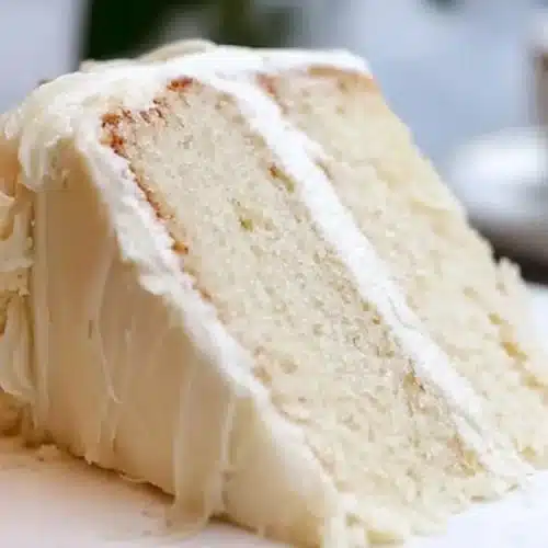 Classic White Almond Cake: The "Mistake-Proof" Bakery Secret for Milestone Memories White Almond Cake A generous slice of moist white almond cake with thick, rustic layers of creamy frosting, served on a classic plate with a cup of coffee resting in the warm background.