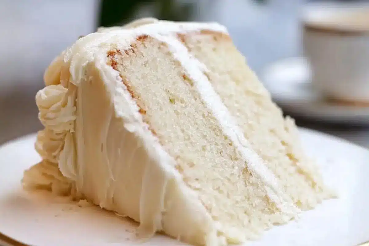 Classic White Almond Cake: The "Mistake-Proof" Bakery Secret for Milestone Memories White Almond Cake A generous slice of moist white almond cake with thick, rustic layers of creamy frosting, served on a classic plate with a cup of coffee resting in the warm background.