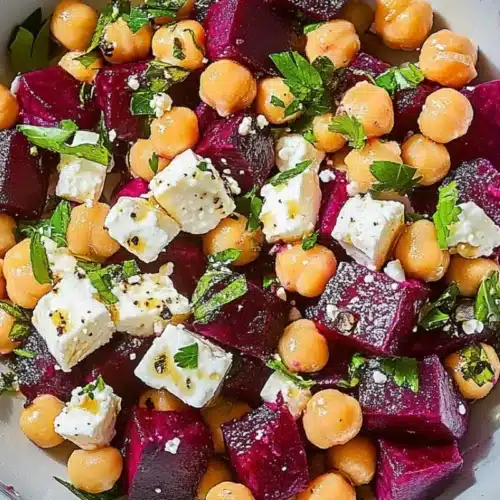 Nonna's Reset Beet and Chickpea Salad with Feta (A Wilt-Proof Dense Bean Bowl) beet and chickpea salad with feta A close-up view of a vibrant beet and chickpea salad with feta, tossed with fresh chopped herbs, black pepper, and a light olive oil dressing in a rustic grey bowl.
