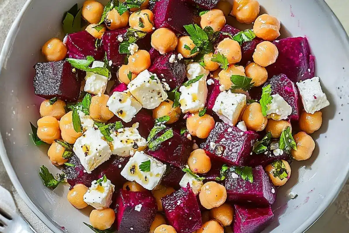 Nonna's Reset Beet and Chickpea Salad with Feta (A Wilt-Proof Dense Bean Bowl) beet and chickpea salad with feta A close-up view of a vibrant beet and chickpea salad with feta, tossed with fresh chopped herbs, black pepper, and a light olive oil dressing in a rustic grey bowl.