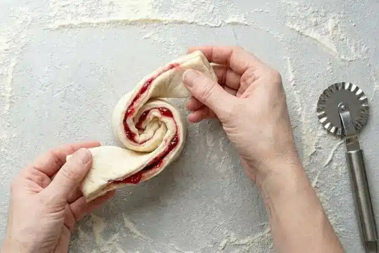 cutting-strips-for-raspberry-pastry-twists Hands twisting a strip of jam-filled puff pastry into shape for Raspberry Pastry Twists on a floured surface.