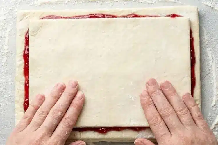 layering-puff-pastry-for-raspberry-pastry-twists (1) Hands placing a second sheet of puff pastry over raspberry jam-covered pastry while assembling Raspberry Pastry Twists on a floured surface.