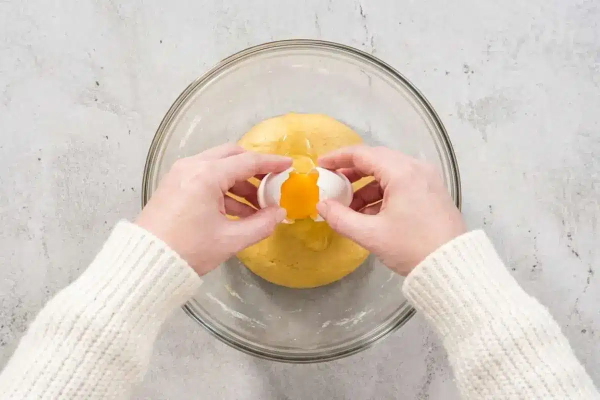 moms-famous-cream-puffs-recipe-adding-eggs (1) A fresh egg being added to warm choux dough in a glass bowl while making Mom’s famous cream puffs recipe.