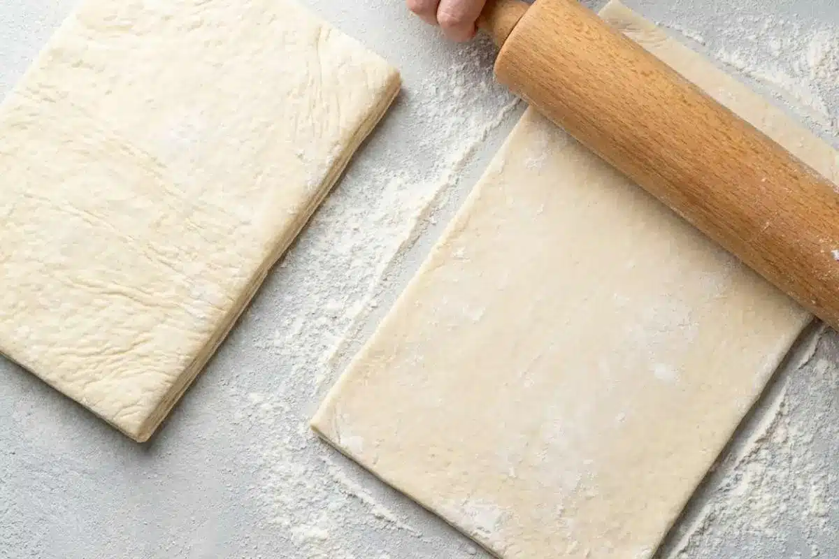 rolling-puff-pastry-for-raspberry-pastry-twists A wooden rolling pin flattening a sheet of puff pastry on a floured surface while preparing Raspberry Pastry Twists.