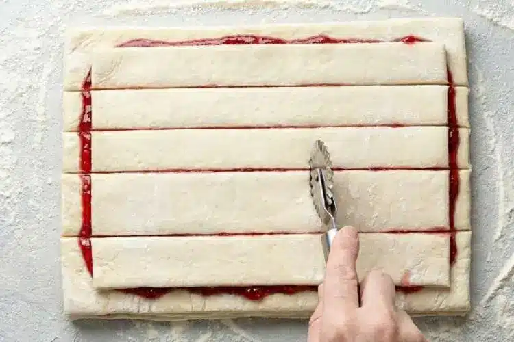 twisting-raspberry-pastry-twists-before-baking A pastry cutter slicing layered puff pastry filled with raspberry jam into strips for Raspberry Pastry Twists on a floured surface
