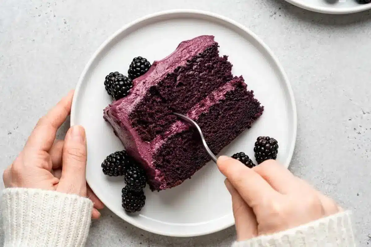 Dreamiest Blackberry Velvet Cake A hand in a cozy white knit sweater uses a silver fork to take a decadent bite from a rich slice of blackberry velvet cake, topped with purple frosting and fresh blackberries on a simple white plate.