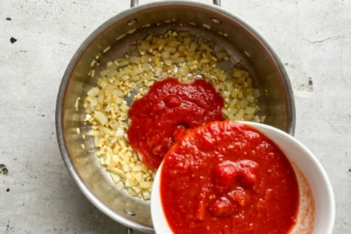 adding-tomatoes-to-authentic-italian-sunday-sauce Crushed tomatoes being poured into a pot with sautéed onions and garlic during the preparation of authentic Italian Sunday sauce.
