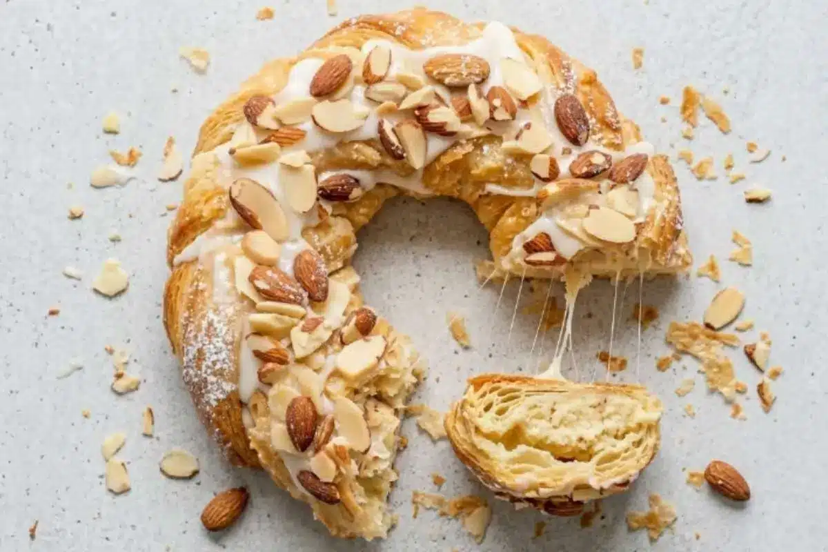 almond-kringle-slice A sliced Almond Kringle showing its flaky layers and creamy almond filling, topped with sweet glaze and toasted almonds on a rustic baking surface.