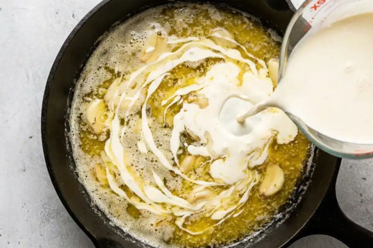 comforting-garlic-parmesan-pasta-cream-sauce (1) Heavy cream being poured into a garlic butter sauce in a skillet for The Most Comforting Garlic Parmesan Pasta recipe.