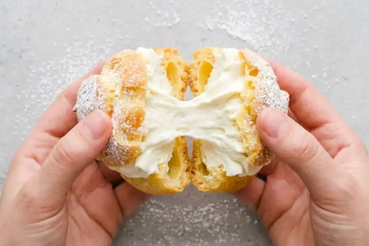 cream-puff-filling-stretchy-cream-center Hands pulling apart a homemade cream puff filled with fluffy cream puff filling and dusted with powdered sugar.