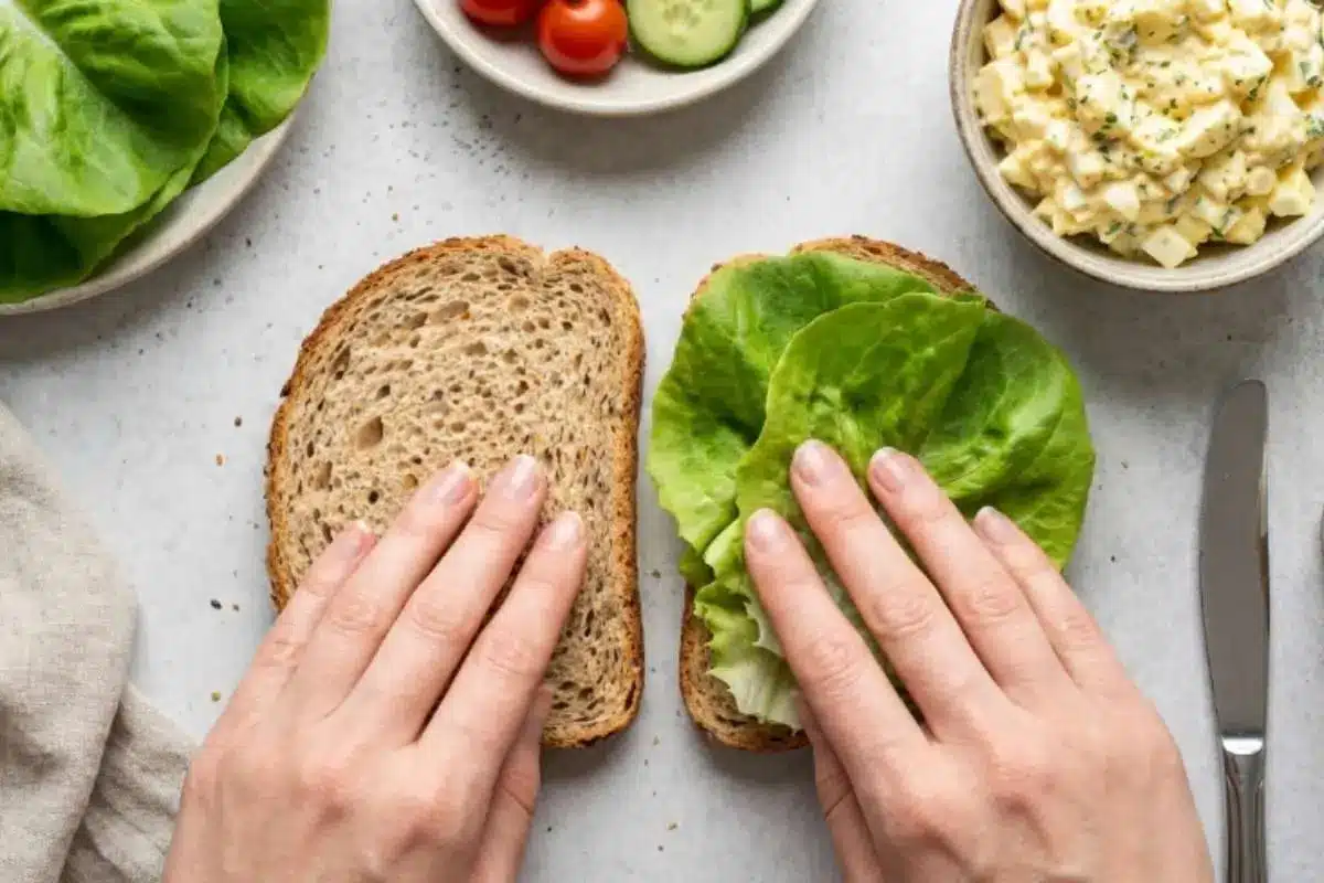 delicious-egg-salad-sandwich-assembly Hands layering fresh lettuce onto whole grain bread while assembling a Delicious Egg Salad sandwich.
