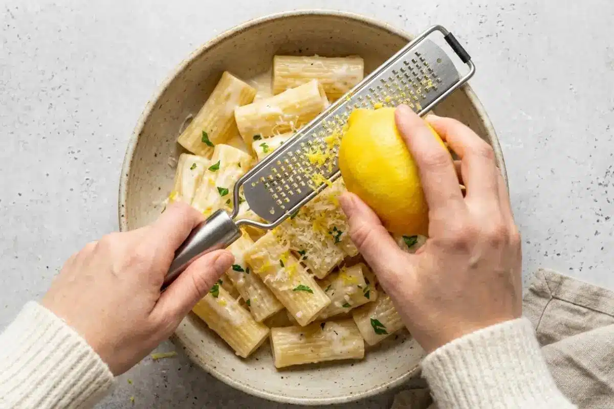 pasta dinner recipes A top-down view of two hands zesting a fresh yellow lemon with a metal grater over a rustic ceramic bowl filled with warm creamy lemon pasta, garnished with grated cheese and fresh parsley.