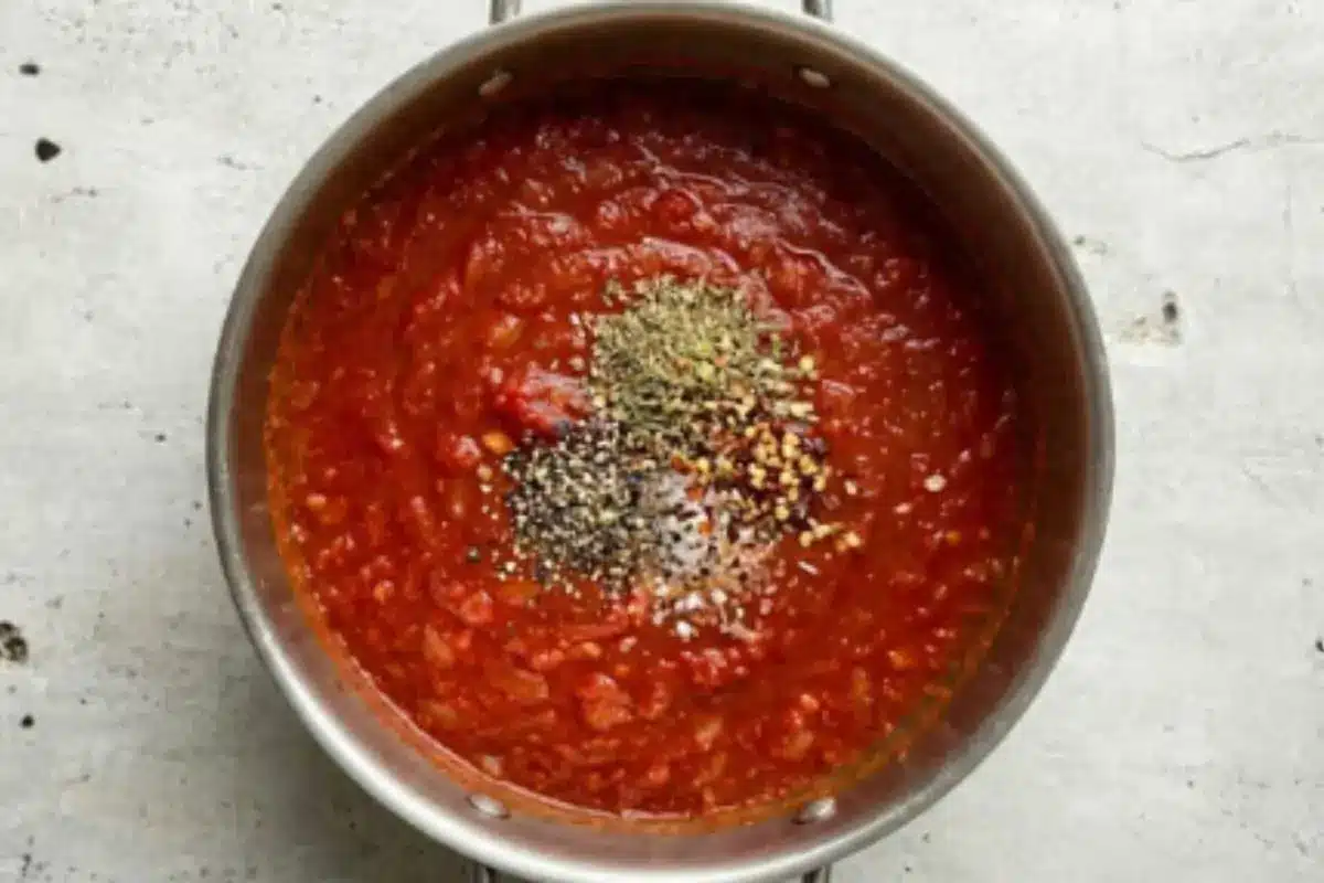 seasoning-authentic-italian-sunday-sauce A pot of authentic Italian Sunday sauce with oregano, red pepper flakes, salt, and black pepper added on top before simmering.
