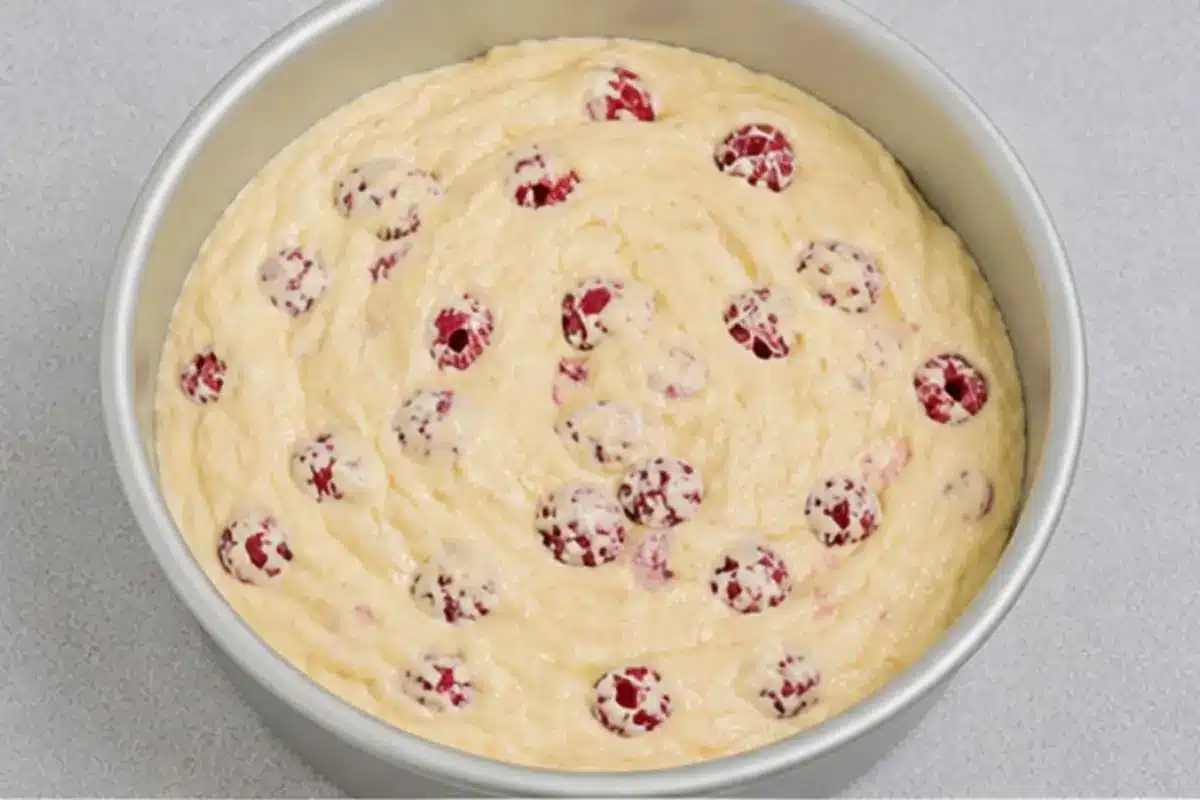 white-chocolate-raspberry-cake-batter-in-pan White chocolate raspberry cake batter spread evenly in a round cake pan, dotted with raspberries before baking.