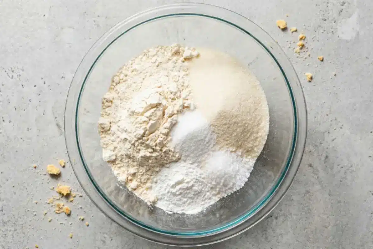 white-chocolate-raspberry-cake-dry-ingredients A glass mixing bowl filled with dry ingredients for white chocolate raspberry cake, including flour, sugar, and baking powder, on a light kitchen surface.