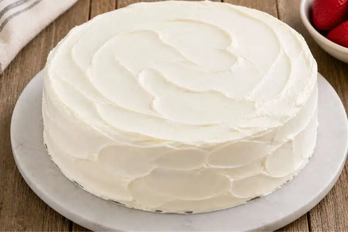 white-chocolate-raspberry-cake-frosted A fully frosted white chocolate raspberry cake with smooth swirls of creamy white frosting on a cake stand over a rustic wooden table.