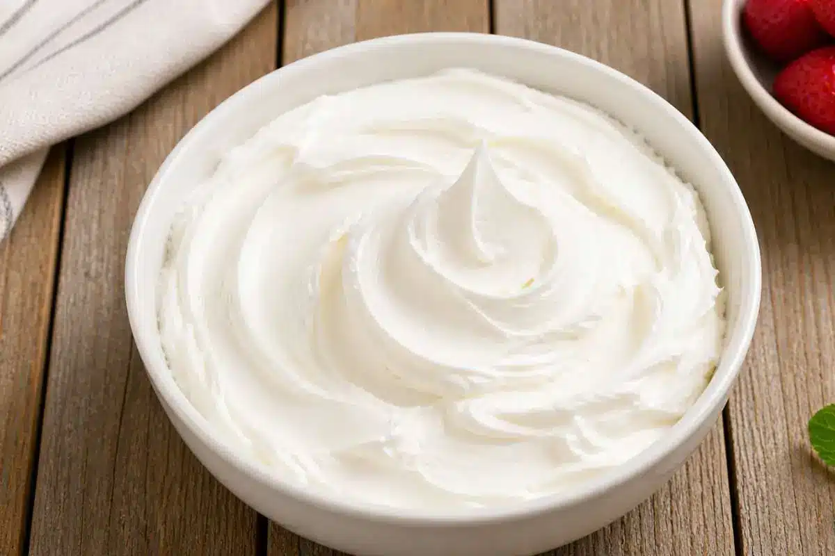 white-chocolate-raspberry-cake-frosting A bowl of smooth and creamy white chocolate frosting for white chocolate raspberry cake, swirled to a soft peak on a rustic wooden surface.