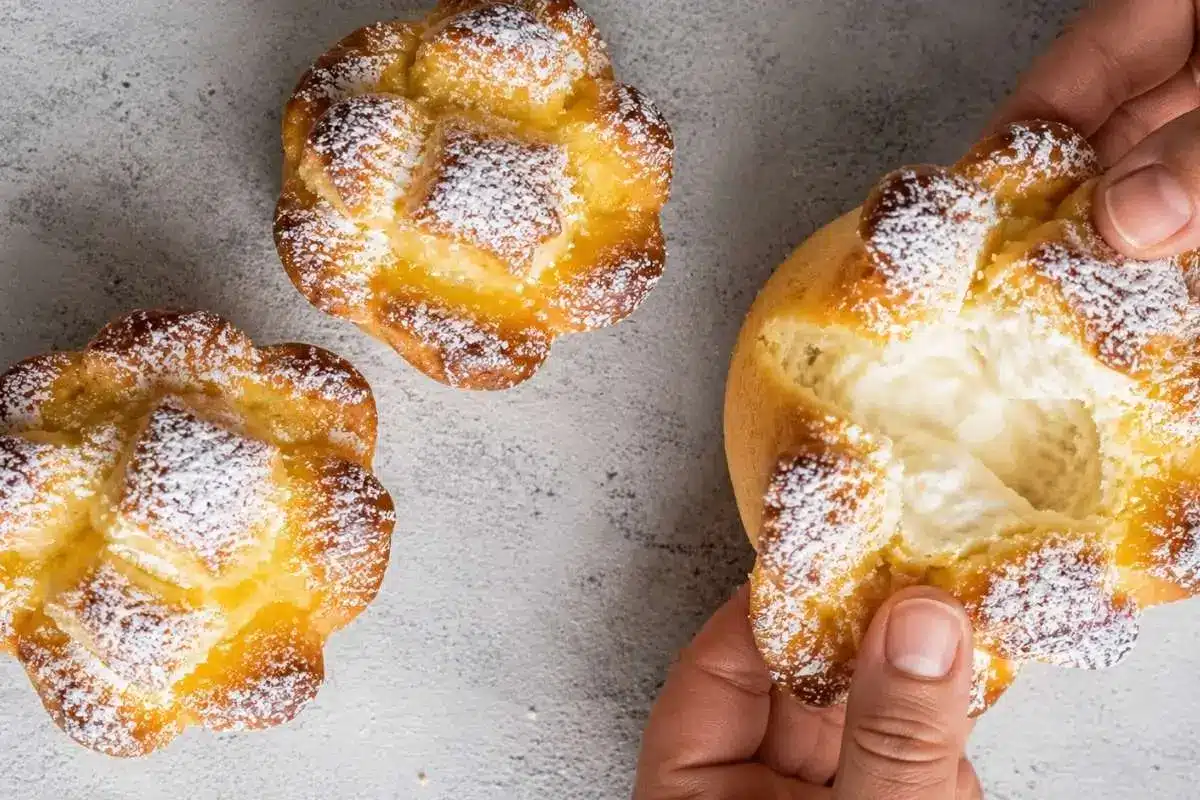 authentic-soffioni-abruzzesi-ricotta-filling A freshly baked Soffioni Abruzzesi sweet ricotta pastry being opened by hand to reveal its airy, creamy ricotta filling, with two powdered sugar-dusted pastries nearby.